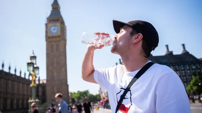 ロンドン・ウェストミンスター橋で、白いTシャツに黒いキャップ姿の男性が、ペットボトルの水を飲んでいる。背景に、ビッグベンや議事堂が並ぶ。