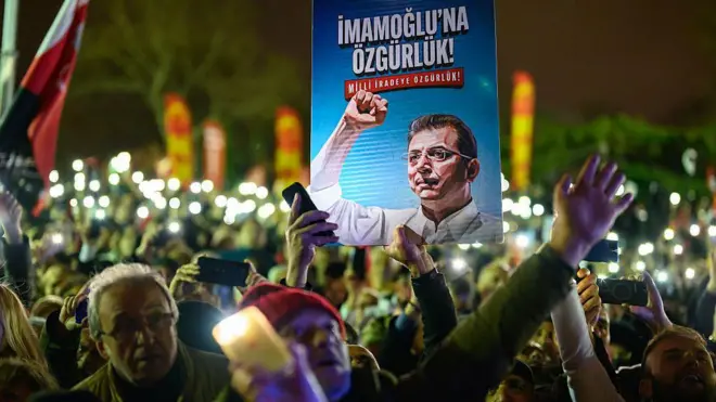 İmamoğlu'nun tutuklanmasının birinci yıl dönümünde düzenlenen gösteride bir protestocu 'İmamoğlu'na özgürlük' yazılı bir poster tutuyor, göstericiler ellerini yukarı kaldırmış ve arka planda telefon ışıkları görülüyor