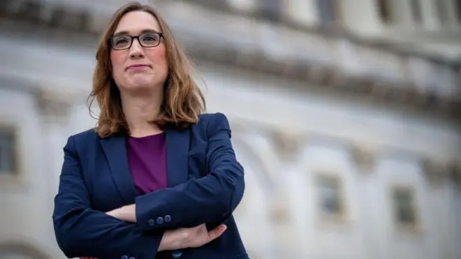 Sarah McBride posando frente al Congreso de EE.UU. Lleva una chaqueta azul, camisa morada, anteojos y pelo largo hasta las mejillas.