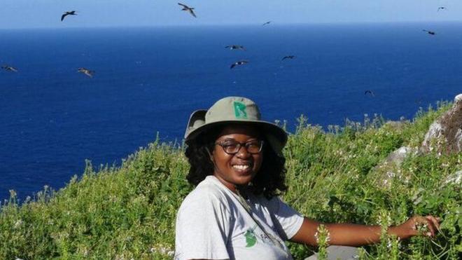 Una ambientalista sentada cerca de una zona verde y detrás se ve el mar y aves volando.