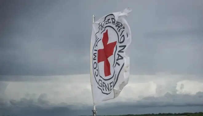 Ibendera ry'umweru rya CICR ririmo umusaraba utukura hamwe n'amagambo yandikishijwe ibara ry'umukara. Inyuma kure haraboneka ibicu by'ubururu n'umweru.