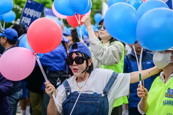 An older female supporter of South Korean presidential candidate Lee Jae-myung of the Democratic Party stands in a crowd with red and pink balloons in hand, wearing sunglasses and a dungaree with a white t-shirt, and smiling.
