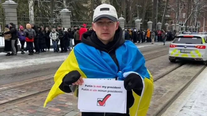 Роман Голиков стоит на улице с флагом Украины на плечах и плакатом с надписью «Putin is not my president»