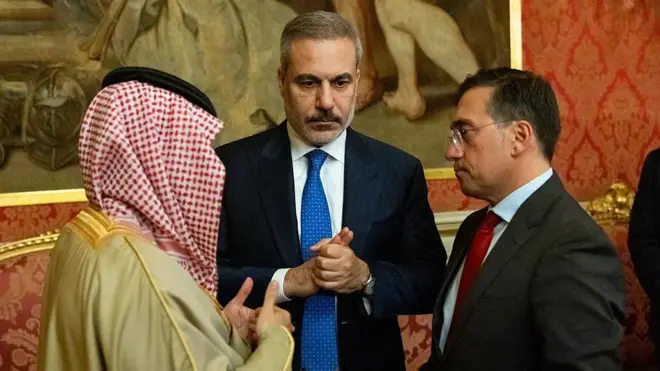 Suudi yerel giysileri içinde bir erkek ile iki takım elbiseli siyasetçi konuşuyor