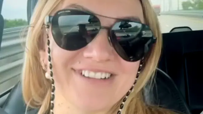 Polina Azarnykh looking straight at the camera while seated in a vehicle. She has long blonde hair and is wearing large sunglasses and a black top and is smiling.