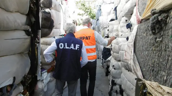 OLAF ve ARPAT yazılı yetkililer tekstik atıklarının arasından geçiyor, arkaları dönük. 