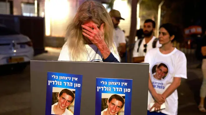 امرأة إسرائيلية تحمل صورتين للملازم هدار غولدين، تبكي وهي تنتظر وصول الجثة التي يُعتقد أنها لغولدين، خارج المركز الوطني للطب الشرعي في تل أبيب. الصورة التقطت ليلاً. خلف المرأة عدد قليل من الأشخاص.