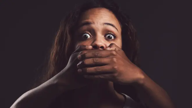 Shot of a scared young woman’s mouth being covered by an unrecognisable man’s hand against a black background