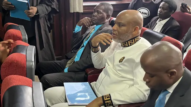 Nnamdi Kanu in court putting on a milky-white Fendi sweater, flanked by two plain-clothe security operatives