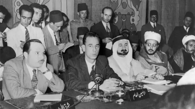 أول أمين عام لجامعة الدول العربية عبد الرحمن عزام باشا خلف المايكروفون، وإلى يمينه ممثل شرق الأردن، وإلى يساره ممثل المملكة العربية السعودية خلال جلسة استثنائية لمناقشة القضية الفلسطينية في سوريا، في 19 يونيو 1946.