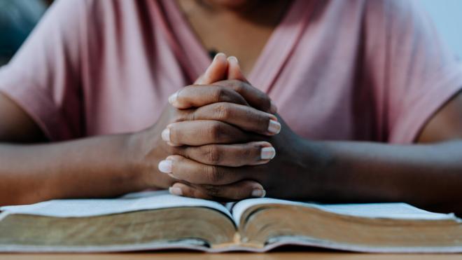 Fotografia mostra mãossportbeting comuma mulher negra entrelaçadas sobre um livro como quem faz uma oração