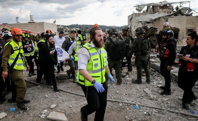Çok sayıda İsrail'li acil durum personeli bir cesedi taşırken görülüyor. Arkada yıkıntılar görünüyor.
