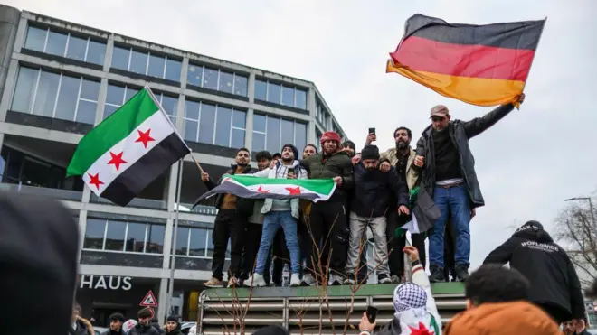 Berlin'de Esad'ın gidişini kutlayan Suriyeli bir grup adam, o dönemki Suriye muhalefetinin bayrağını ve Alman bayrağını taşıyor.