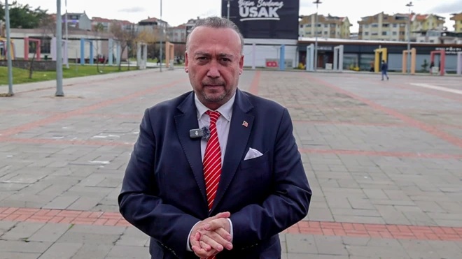 Özkan Yalım kent meydanında ayakta duruyor, ellerini birbirine bağlamış, kameraya bakıyor. 