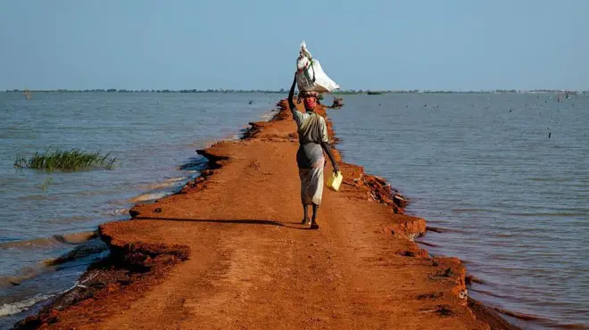 एक महिला अपने सिर पर भारी बोझ उठाए हुए पतली ज़ीरो-किलो 30 सड़क पर चल रही है, जो बेंटियू, यूनिटी स्टेट, दक्षिण सूडान के पास है