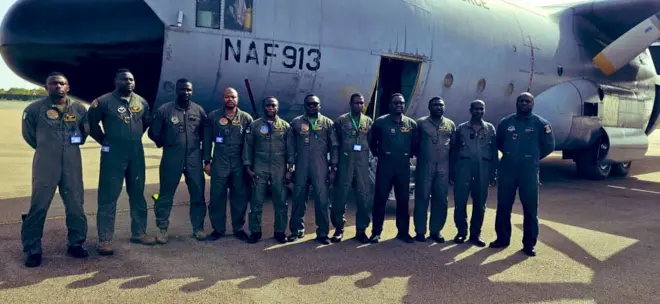 Àwọn ọmọ ogun mọ́kànlá tó gba ìtúsílẹ̀ dúró sí iwájú ọkọ̀ òfurufú