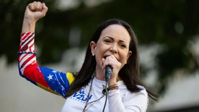 María Corina Machado azamuye igipfunsi arimo kuvugira mu ndangururamajwi. Yambaye umupira w'amaboko maremare urimo amabara y'ubururu, umweru, umuhondo n'umutuku y'ibendera rya Venezuela.