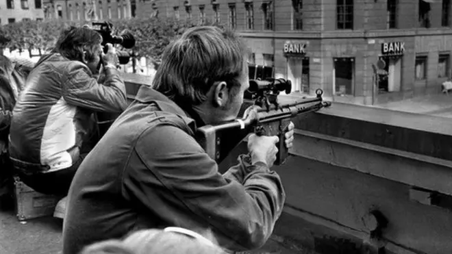 Two policemen hold guns, poised opposite a Kreditbanken building