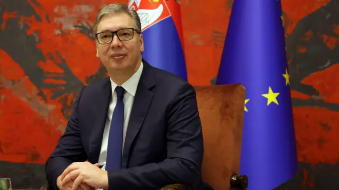 Председник Вучић у оделу седи склопљених руку са заставама Србије и Еу иза леђа