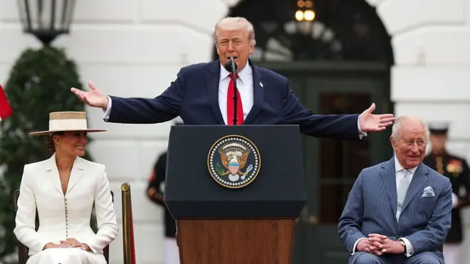 Donald Trump stoi przy mównicy prezydenta Stanów Zjednoczonych, mając uniesione obie ręce na boki. Po jednej stronie siedzi król Karol, po drugiej żona Trumpa, Melania. Król ma na sobie jasnoniebieski garnitur w delikatne paski, jasną koszulę oraz jasnoniebieski krawat z pasującą poszetką; patrzy w bok i śmieje się. Trump ubrany jest w granatową marynarkę, białą koszulę i czerwoną krawatkę, przemawiając do zgromadzonych. Melania ma na sobie dopasowany biały garnitur oraz słomkowy kapelusz. Scena rozgrywa się przed Białym Domem.