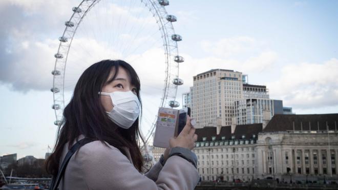 一名戴上口罩的亞洲女子在倫敦西敏寺橋上拿著手機朝倫敦眼摩天輪拍照（2/3/2020）