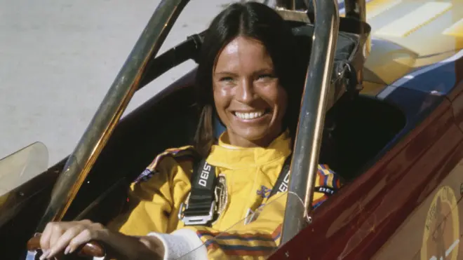 基蒂·奧尼爾(Kitty O'Neil)——一位留著烏黑直髮的女性——坐在一輛敞篷車中,身穿黃色連身衣,雙手握著方向盤,對著鏡頭露出微笑(7/7/1977)