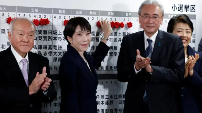 報道陣に向かって笑みを浮かべるダークスーツ姿の首相と拍手する自民党幹部たち