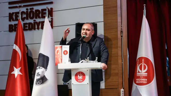 Özarslan bir kürsüde konuşuyor yanında Atatürk'ün kalpaklı görselinin olduğu bayrak onun yanında Türk bayrağı var. 
