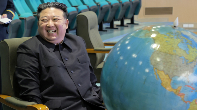 Kim smiles and laughs with subordinates during a meeting with satellite launch committee members. He is sitting at a desk behind a big globe of the world