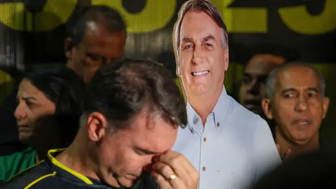 Flávio Bolsonaro com a cabeça baixa, e, no fundo, uma imagem de Bolsonaro impressa em papelão de tamanho real. 