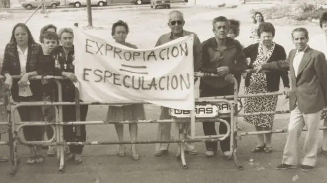 Un grupo de personas, principalmente mayores, están de pie junto a una baranda que parece cerrar el acceso a una calle. En el centro dos de ellos cargan una pancarta que dice "expropiación=especulación".