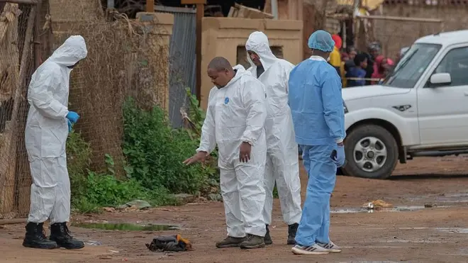 South African Police Service Forensic Pathology Services pipo wey dey wear white and blue overalls dey point for di scene wey di attack happun. Dem dey show one heap of cloth wey dey for di muddy ground.