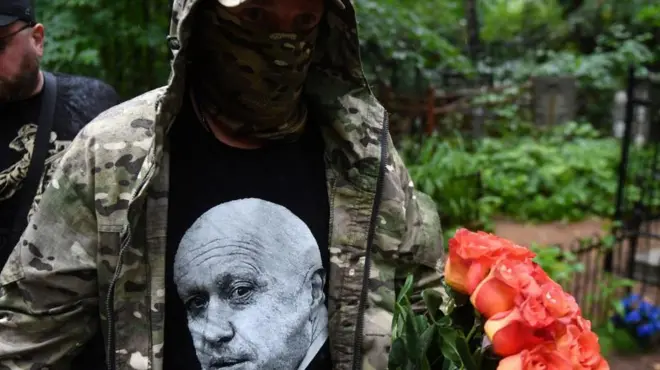 مردی با چهره پوشیده با اورکت نظامی و پیراهنی که تصویر پریگوژین روی آن هست