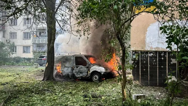 У дворі житлового будинку горить білий фургон: з-під капота і салону виривається вогонь і густий дим. Навколо — повалене листя й уламки, поруч стоять дерева, а на тлі видно багатоповерхівку з пошкодженими вікнами.