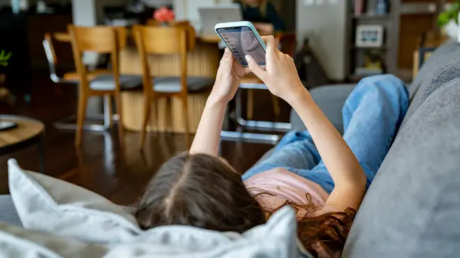 一名少女在家中躺在沙发上滑手机并浏览社交媒体。