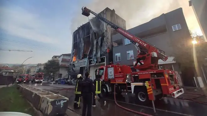 Fotoğrafta yanmış bir bina önünde bir itfaiye aracı var. Aracın önünde üç kişi var. 