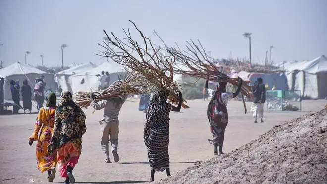 تظهر نساء سودانيات من الخلف باللباس التقليدي يحملن حطباً، وعدد آخر من الرجال يحمل أحدهم حطباً. كل شخص في الصورة يظهر ماشياً لوحده قرب عدد من الخيام. الصورة التقطت نهاراً.