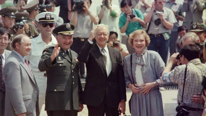 جیمی کارتر، رئیس‌جمهور سابق ایالات متحده پس از عبور از مرز کره شمالی در ژوئن ۱۹۹۴، همراه با همسرش روزالین، برای خبرنگاران دست تکان می‌دهد 
