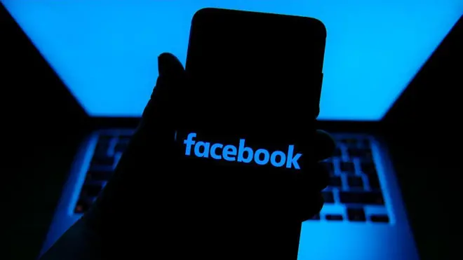 O logotipo do Facebook é exibido na tela de um celular, à frente de um laptop