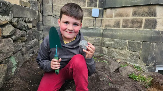A boy, wearing red trousers, a grey hoodie and holding a fossil.