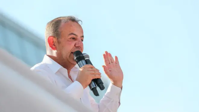 Tanju Özcan üzerinde beyaz bir gömlek ve elinde iki mikrofonla bir konuşma yaparken