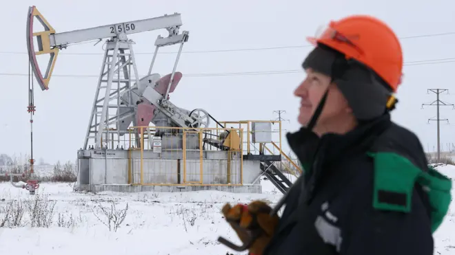 Зимний пейзаж. На заднем плане нефтяной станок-качалка. На переднем справа человек в профиль в рабочей одежде и оранжевой каске.