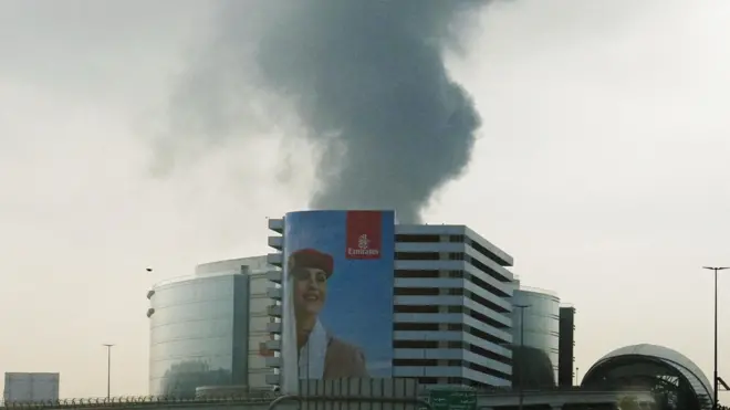 Smoke rising from an area near Dubai International Airport is seen through a vehicle’s windshield after a drone attack hit a fuel tank. In the foreground, an Emirates poster portraying a female flight attendant covers almost the entire façade of a building