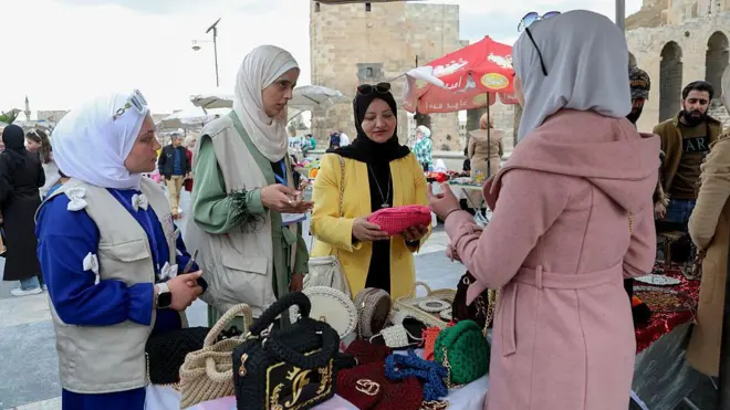 Üç kadın bir tezgahta çantalara bakıyor, pembe paltolu bir kadın onlara ürünleri anlatıyor