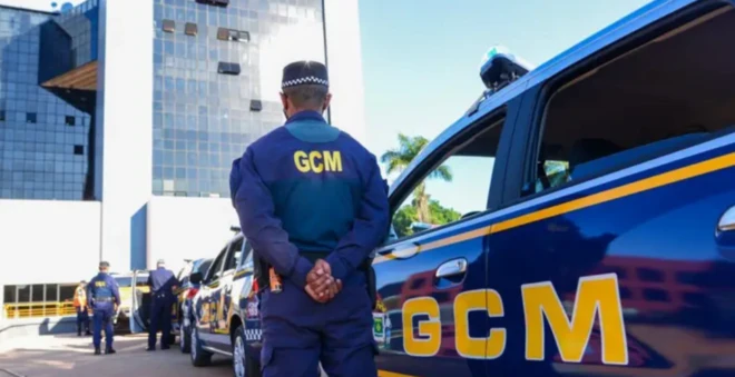 Foto de um guarda civil municipal de Goiânia em frente a uma viatura azul com letras amarelas: GCM
