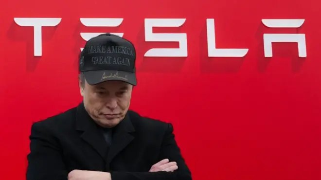 ایلان ماسک در مقابل آرم شرکت تسلا