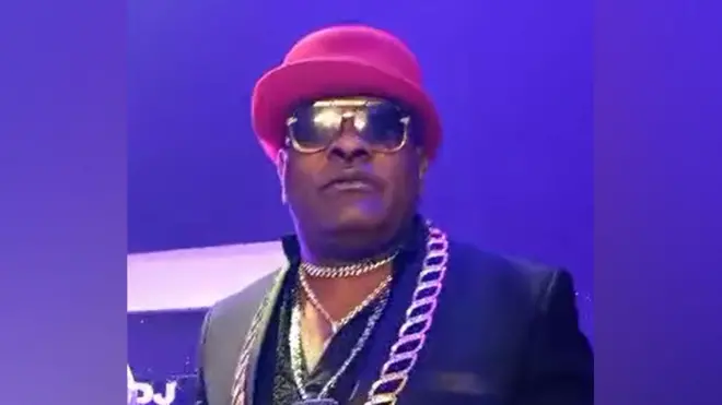 Extrait d'une vidéo de Longue Longue Longue lors d'un concert. Il porte des chaînes en or, des lunettes noires et un chapeau melon rouge.