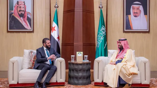 الشرع يلتقي ولي العهد السعودي محمد بن سلمان، يرتدي بن سلمان الزي السعودي التقليدي الرسمي ويصافح الشرع الذي يرتدي بدلة رسمية.