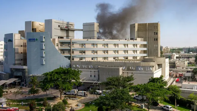 A wide view shows smoke rising from behind the Soroka medical centre against a blue sky and with cars and fire trucks outside it, in Beersheba on Thursday