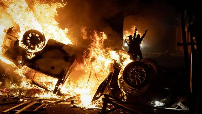 An upturned vehicle blaze sin flames while two silhouetted figures appear to cheer in the background